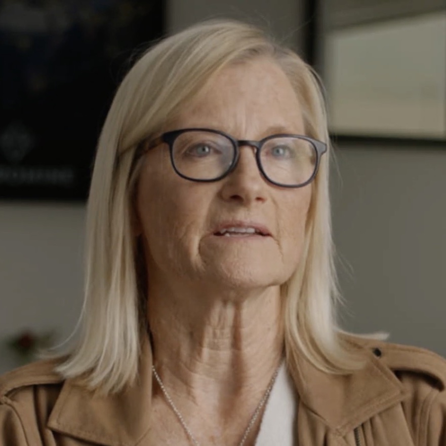 Middle-aged woman with blonde hair and black-rimmed glasses wearing a tan jacket, looking thoughtfully to the side.