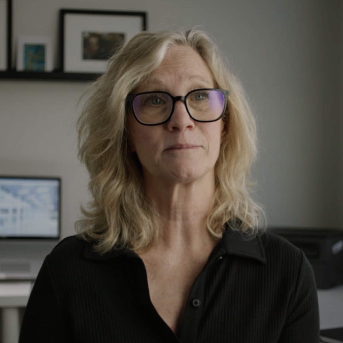 Middle-aged woman with blonde hair and glasses wearing black shirt in modern office with computers and framed artwork.