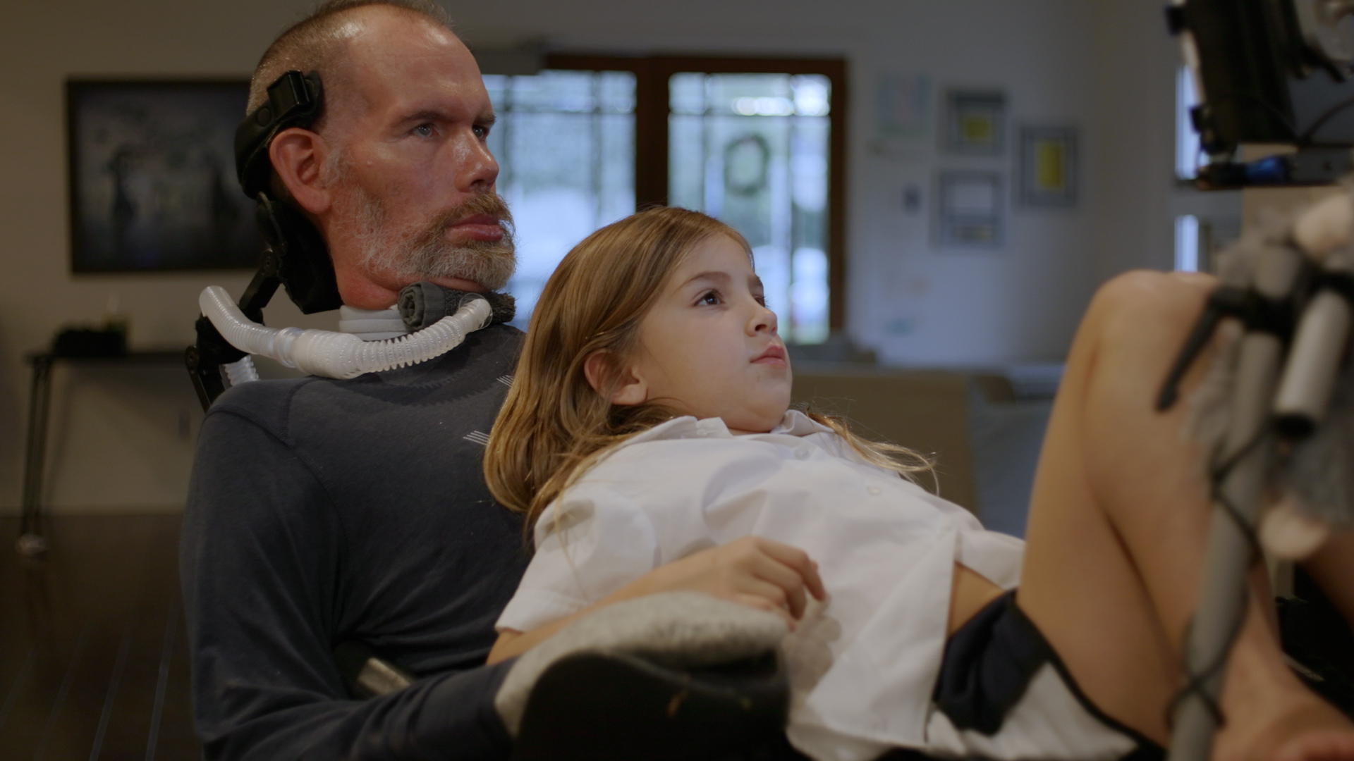 A man using assistive breathing equipment holds a young girl in a white shirt as they both look upward together indoors.