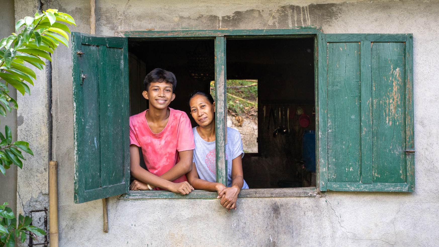 Two smiling people, a teen boy and woman, lean out of a rustic window with weathered green wooden shutters on a concrete wall.