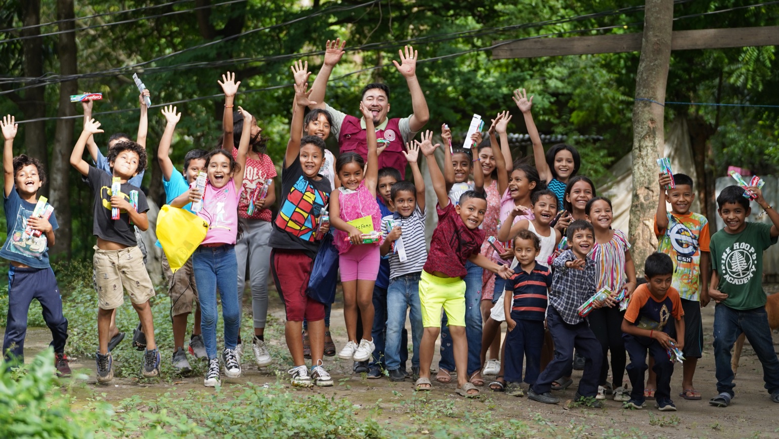 Large group of cheerful children and young adults celebrating together outdoors, raising their hands in joy while holding books and supplies.
