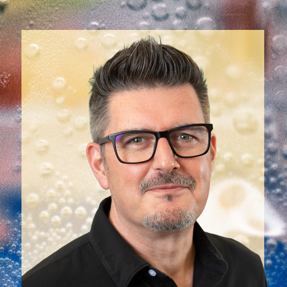 A middle-aged man with dark salt-and-pepper hair, a goatee, and black rectangular glasses wearing a black shirt, posed against a colorful abstract background with bubble-like textures.