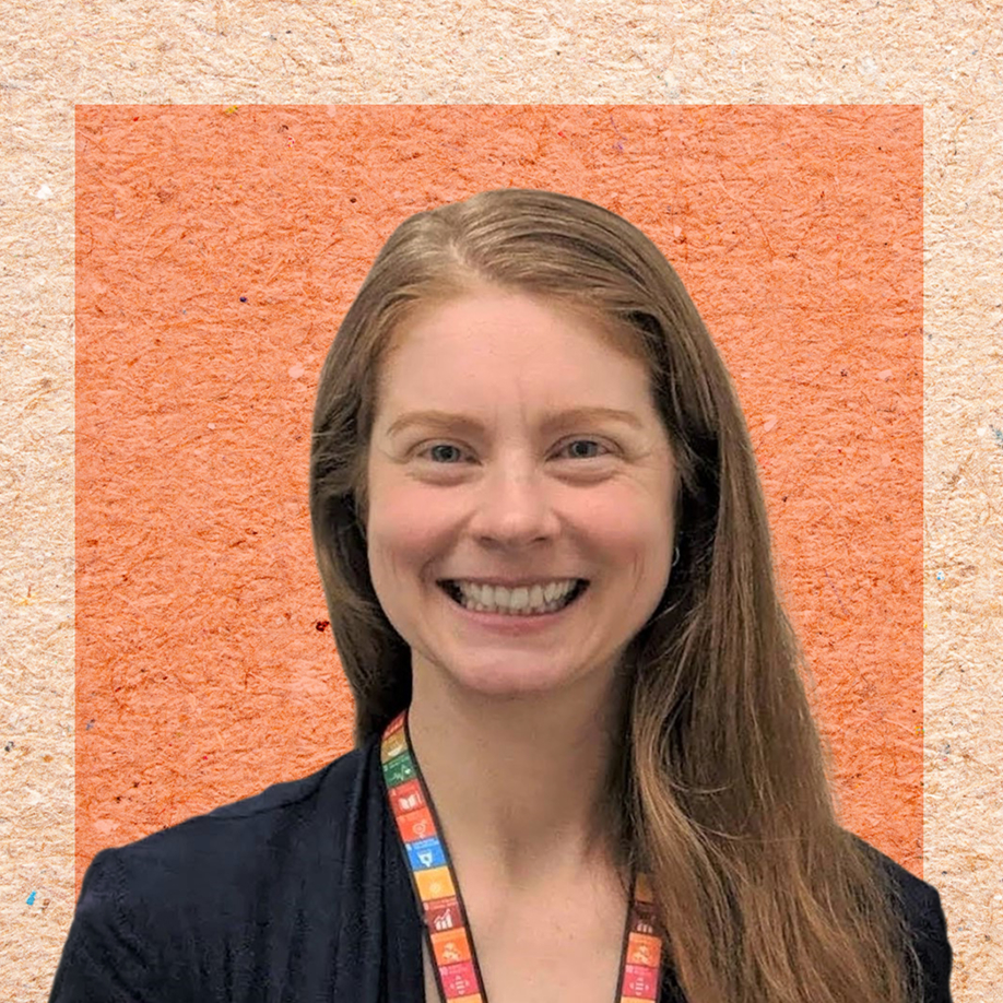A smiling woman with long auburn hair wearing a dark blazer and a colorful UN Sustainable Development Goals lanyard, photographed against an orange textured background.