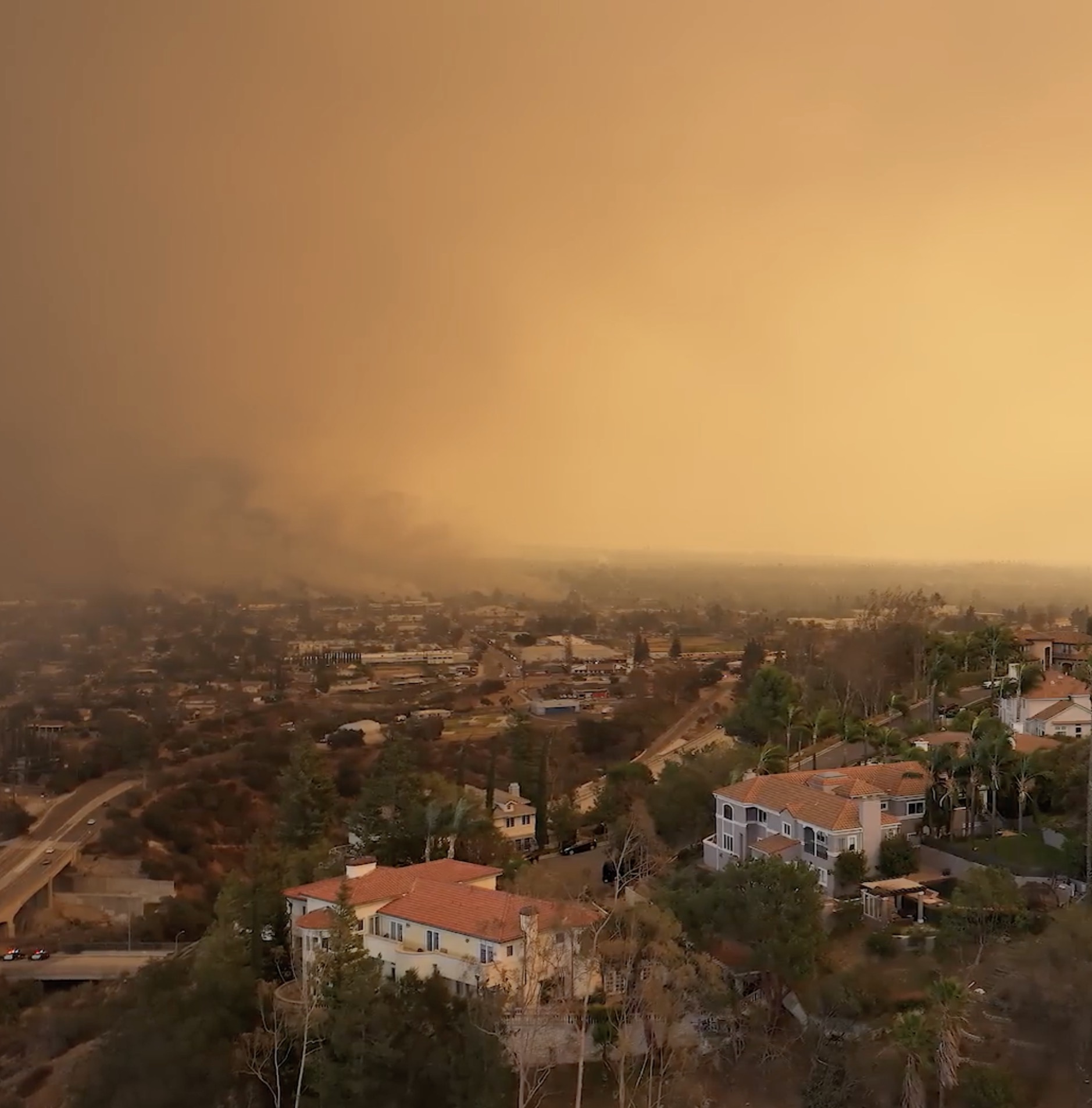 Aerial view of a suburban neighborhood with Mediterranean-style homes and palm trees, blanketed under a thick, orange-tinted haze from a nearby wildfire with dark smoke visible on the horizon.