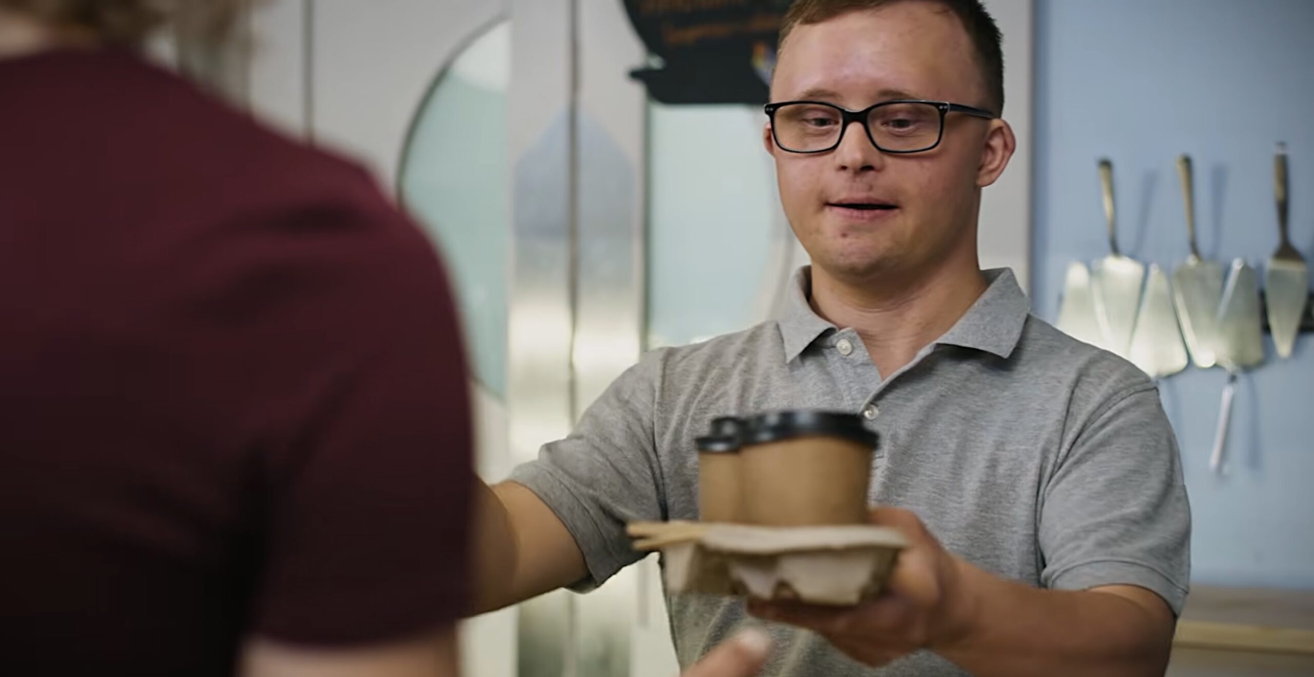 A man in a gray polo shirt hands a takeout coffee cup and napkins to a customer at a counter.