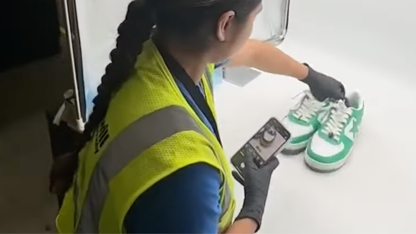 A person in a yellow safety vest and black gloves photographs a pair of green and white sneakers on a white surface.