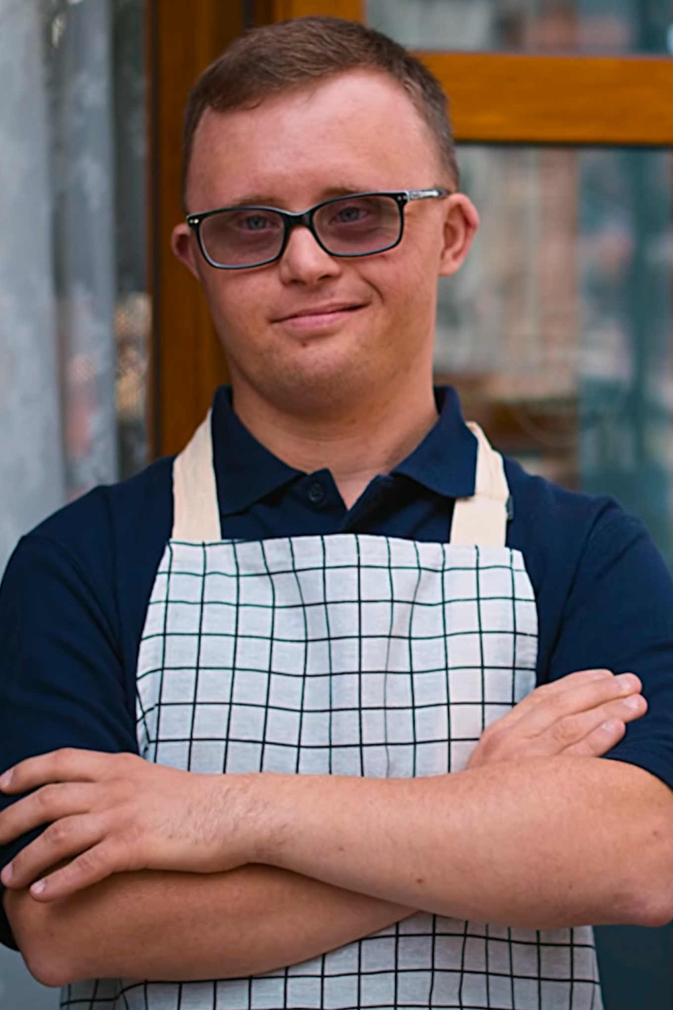 A person wearing glasses and a checkered apron stands outside with arms crossed, smiling at the camera.