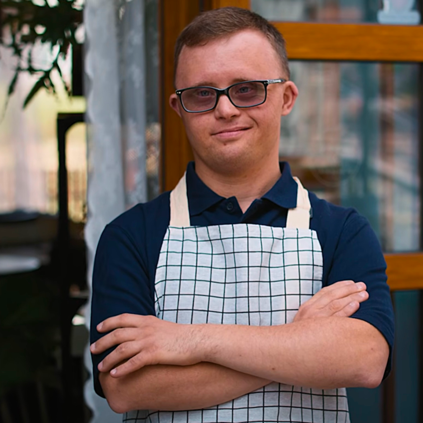 A person wearing glasses and a checkered apron stands outside with arms crossed, smiling at the camera.