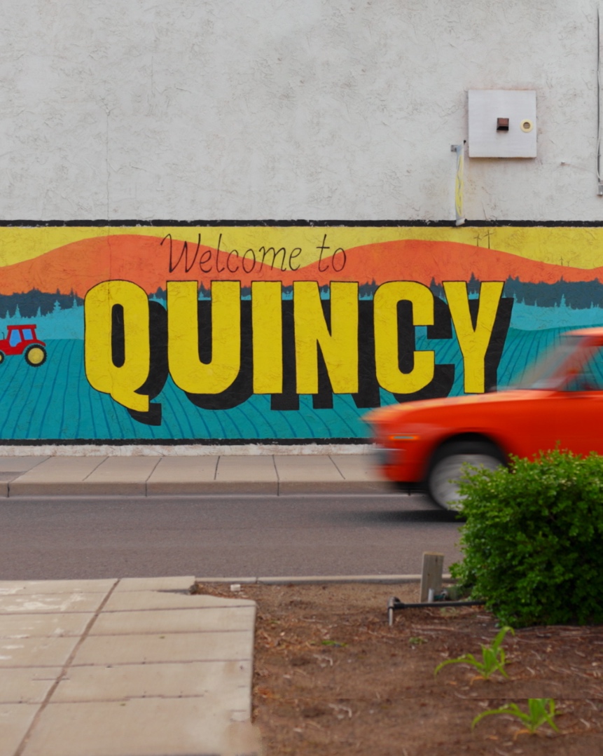 Colorful "Welcome to Quincy" wall mural featuring bold yellow lettering, a teal farmland landscape, red tractor, and orange sunset design, with a motion-blurred