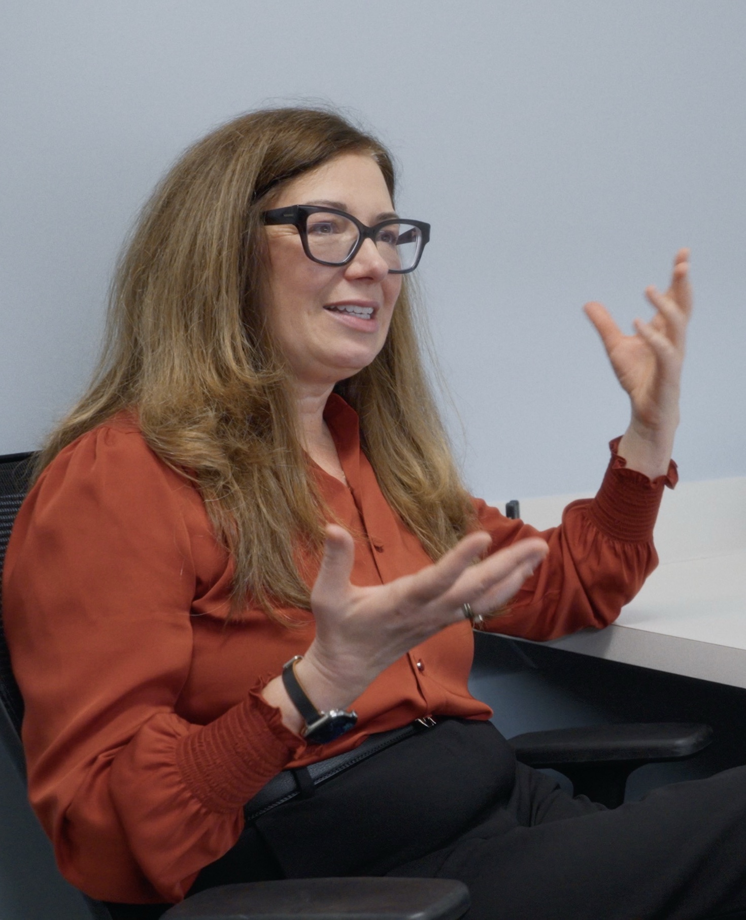 Woman named Andrea seated in an office chair, speaking and gesturing with both hands, wearing a rust-colored blouse, black-rimmed glasses, and a watch