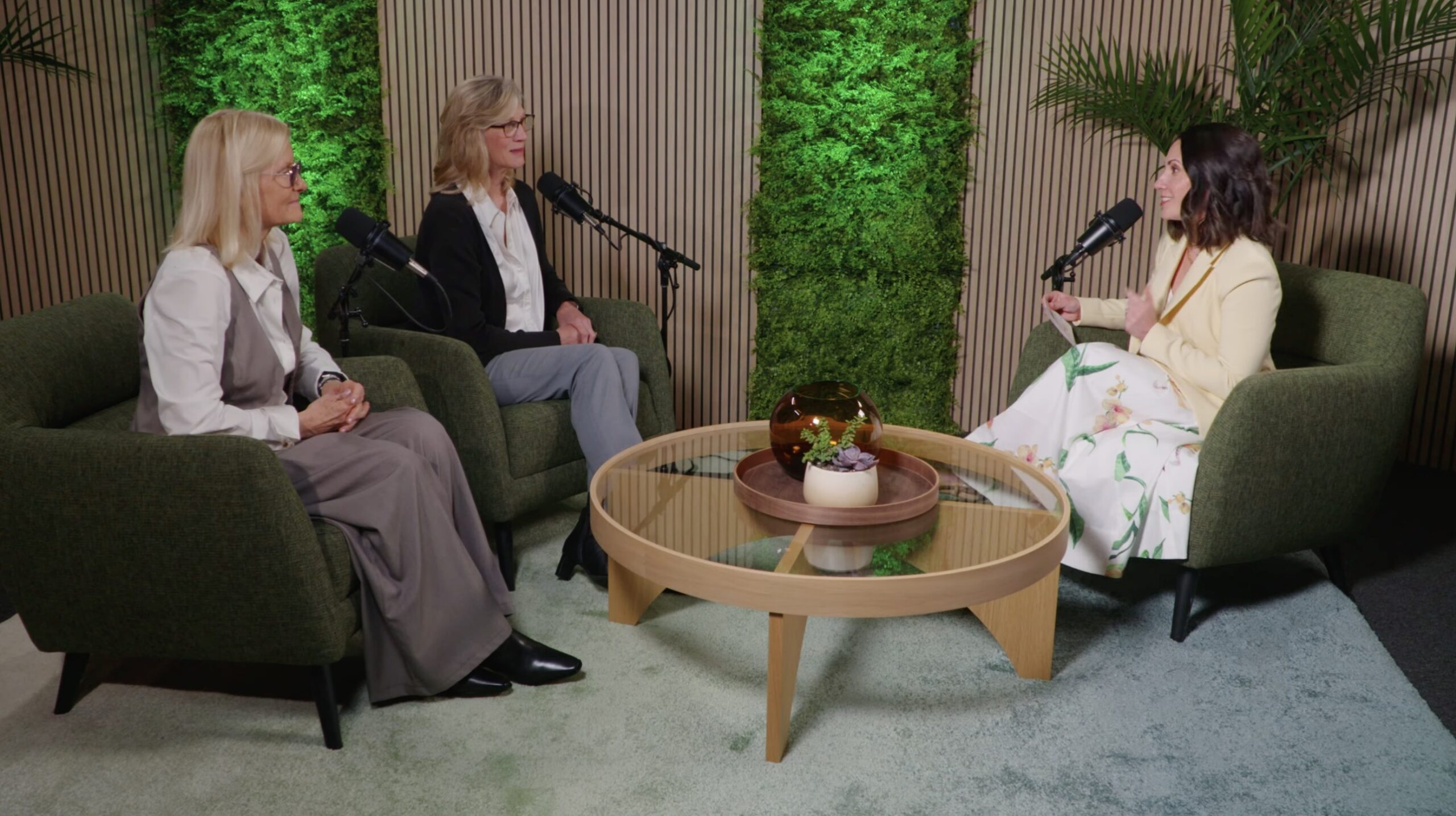 Three women seated in green armchairs during a podcast recording, with microphones positioned between them and a round wooden coffee table at center, set against a