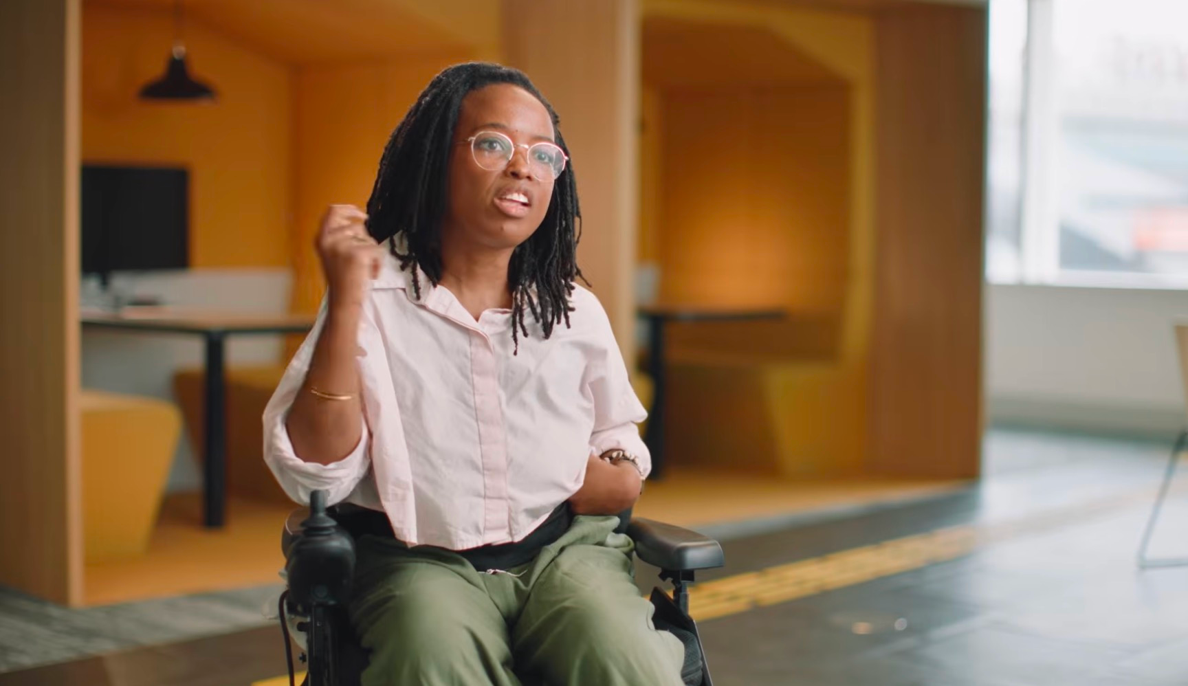 A woman with locs and pink-framed glasses, wearing a light pink button-down shirt and olive green pants, sits in a power wheelchair and speaks expressively with a