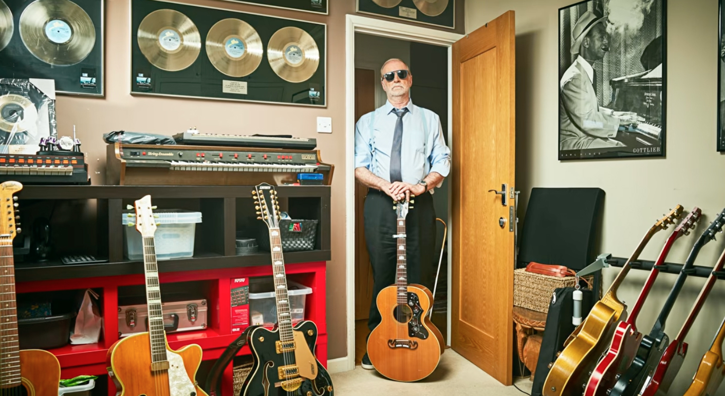 Older man wearing sunglasses, a light blue shirt, and dark tie standing in a doorway surrounded by a large collection of guitars, with gold record awards and a jazz
