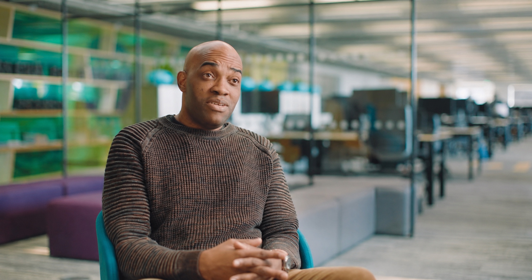 Kwesi, a bald man wearing a brown ribbed sweater, sits in a teal chair with hands clasped, speaking to camera in a modern open-plan office environment