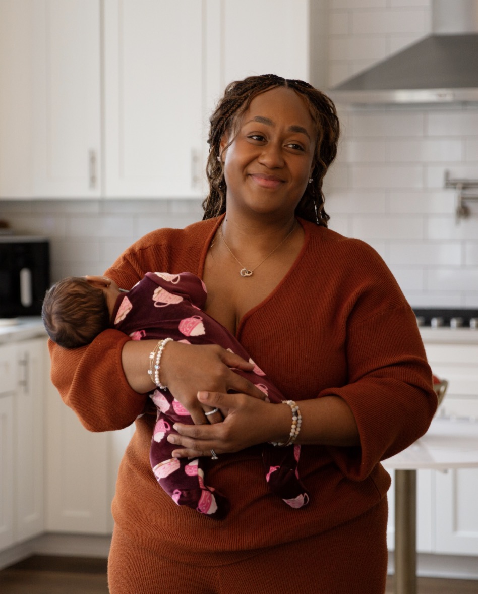 Ashlee smiling and cradling a newborn baby wrapped in a purple and pink printed swaddle, standing in a modern white kitchen wearing an rust-orange sweater and pearl