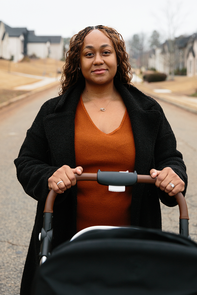 A woman wearing an orange ribbed dress and black teddy coat pushes a black stroller down a residential street on an overcast day