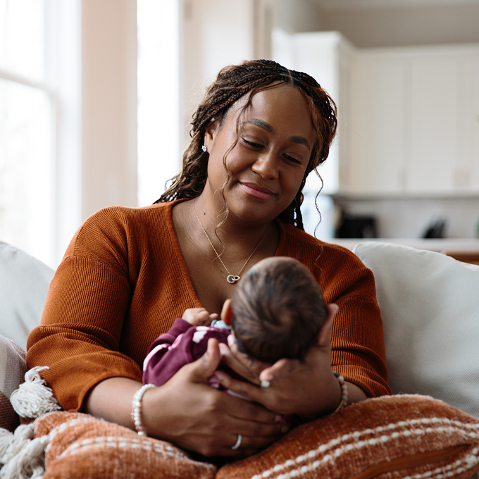 A woman with braided hair wearing an orange sweater sits on a couch at home, smiling warmly as she cradles a newborn baby in her arms