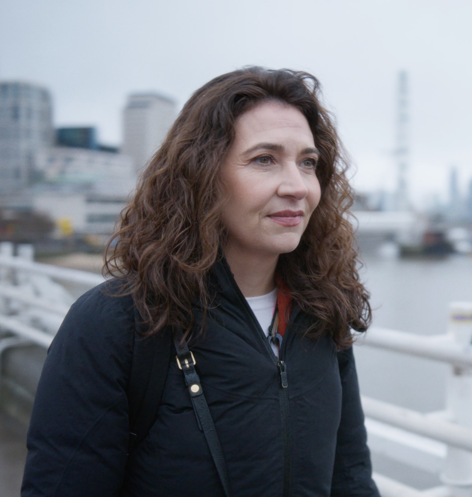 Woman named Paula with curly brown hair, wearing a dark quilted jacket and crossbody bag, standing on a riverside walkway with a blurred urban skyline and river in