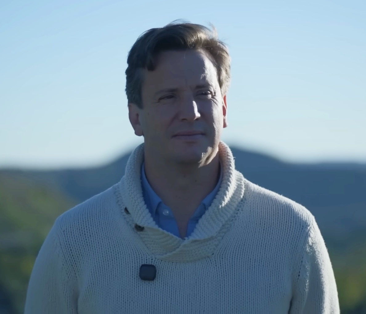 Man wearing a cream shawl-collar sweater over a light blue shirt, standing outdoors with a hilly landscape and clear blue sky in the background