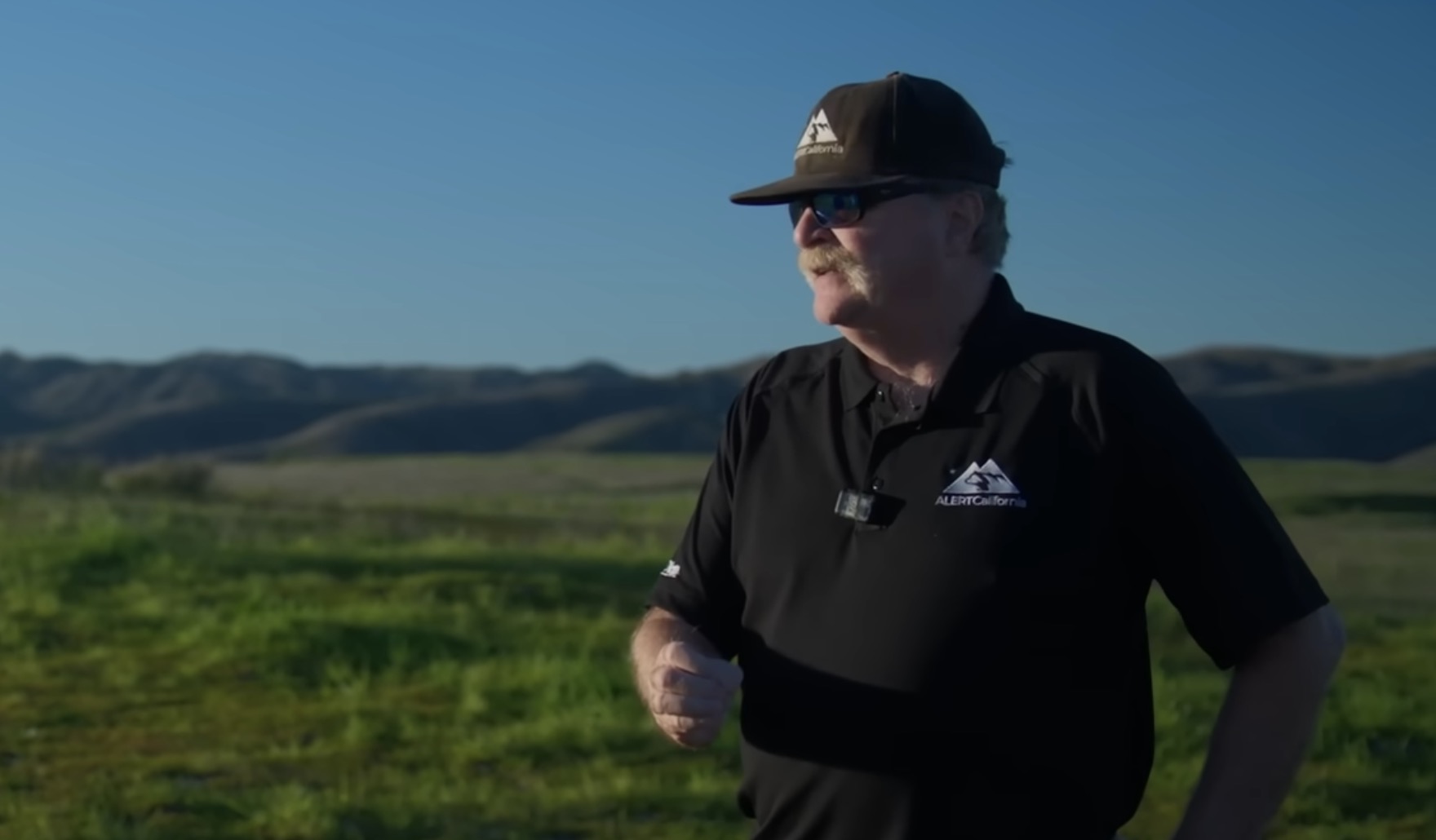 Man wearing an ALERTCalifornia branded black polo shirt and matching cap with sunglasses, standing outdoors in a rural landscape with green fields and hills in the
