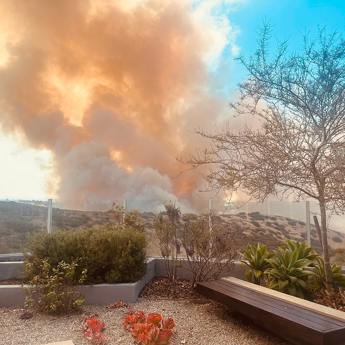 Large plumes of orange and white smoke billowing from a wildfire burning across dry hillside terrain, viewed from a residential outdoor patio with garden planters