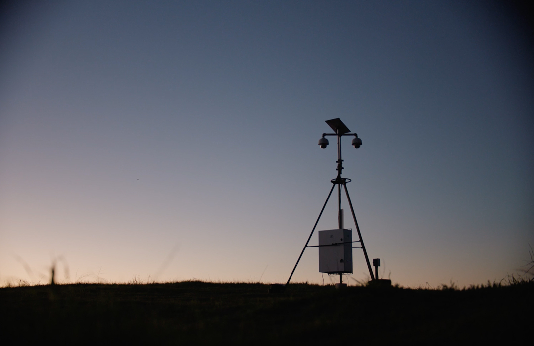 Solar-powered remote surveillance tower with dual PTZ cameras mounted on a tripod mast, silhouetted against a twilight sky over an open field