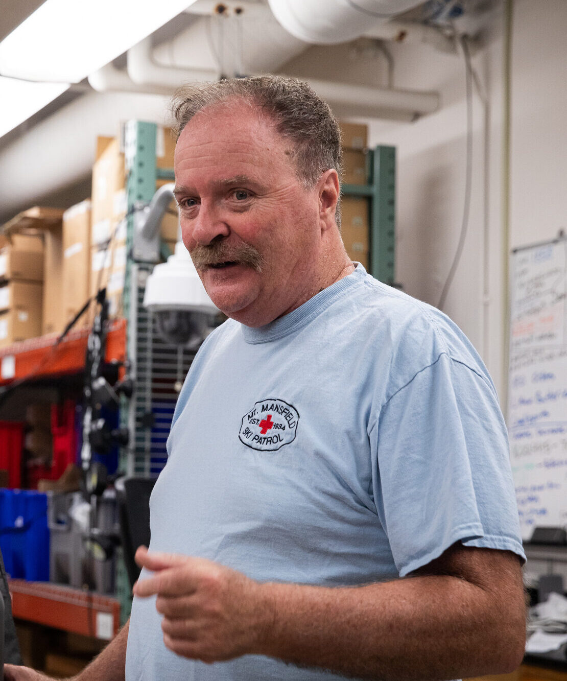 Man with a mustache wearing a light blue Mt. Mansfield Ski Patrol t-shirt, speaking in what appears to be a warehouse or workshop setting with shelving and