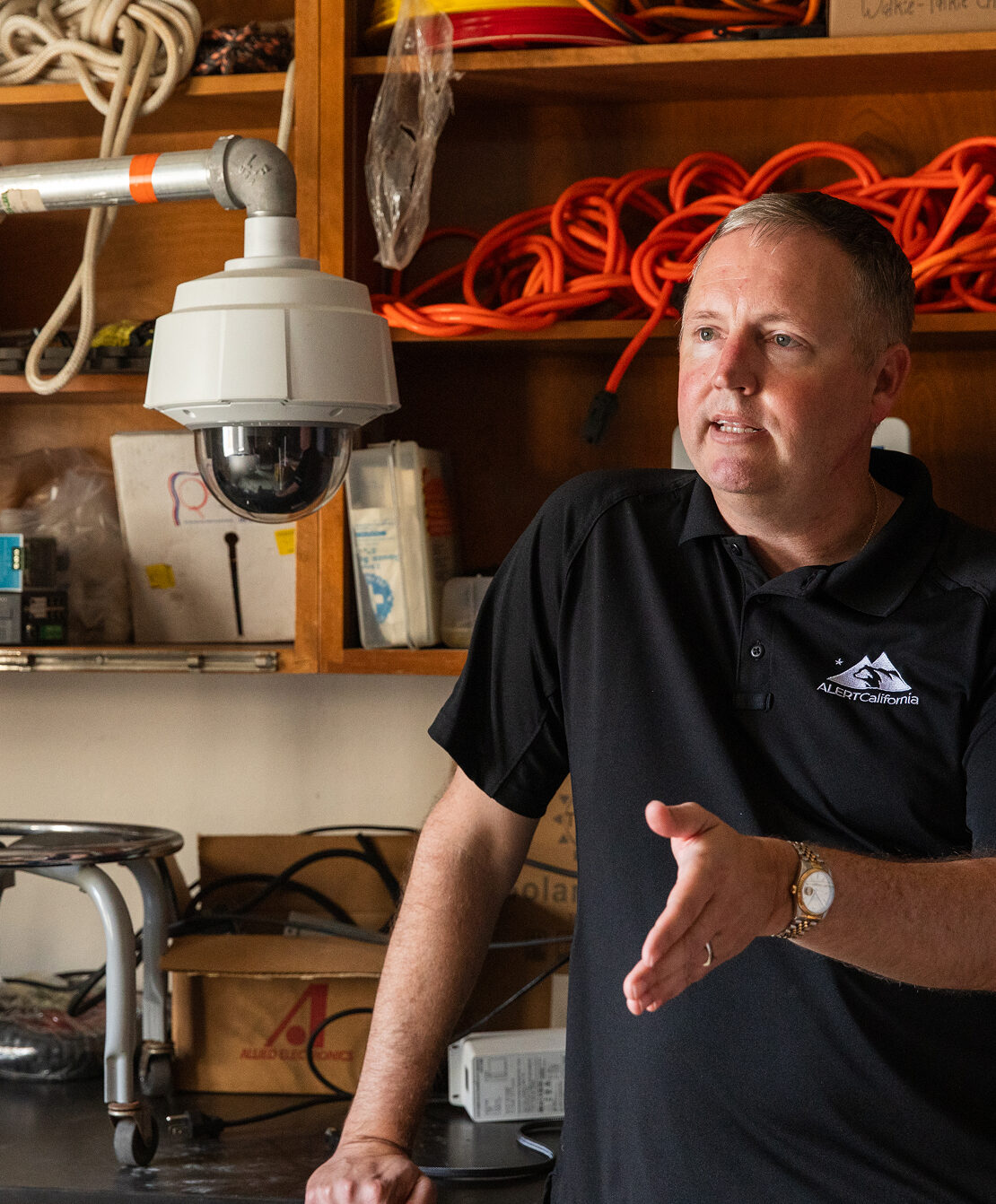 A man wearing a black ALERTCalifornia polo shirt gestures while speaking in a storage room filled with shelves holding orange extension cords, rope, and equipment