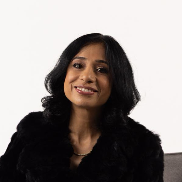 Woman with shoulder-length black hair smiling, wearing a black fur coat and gold necklace, seated against a white background