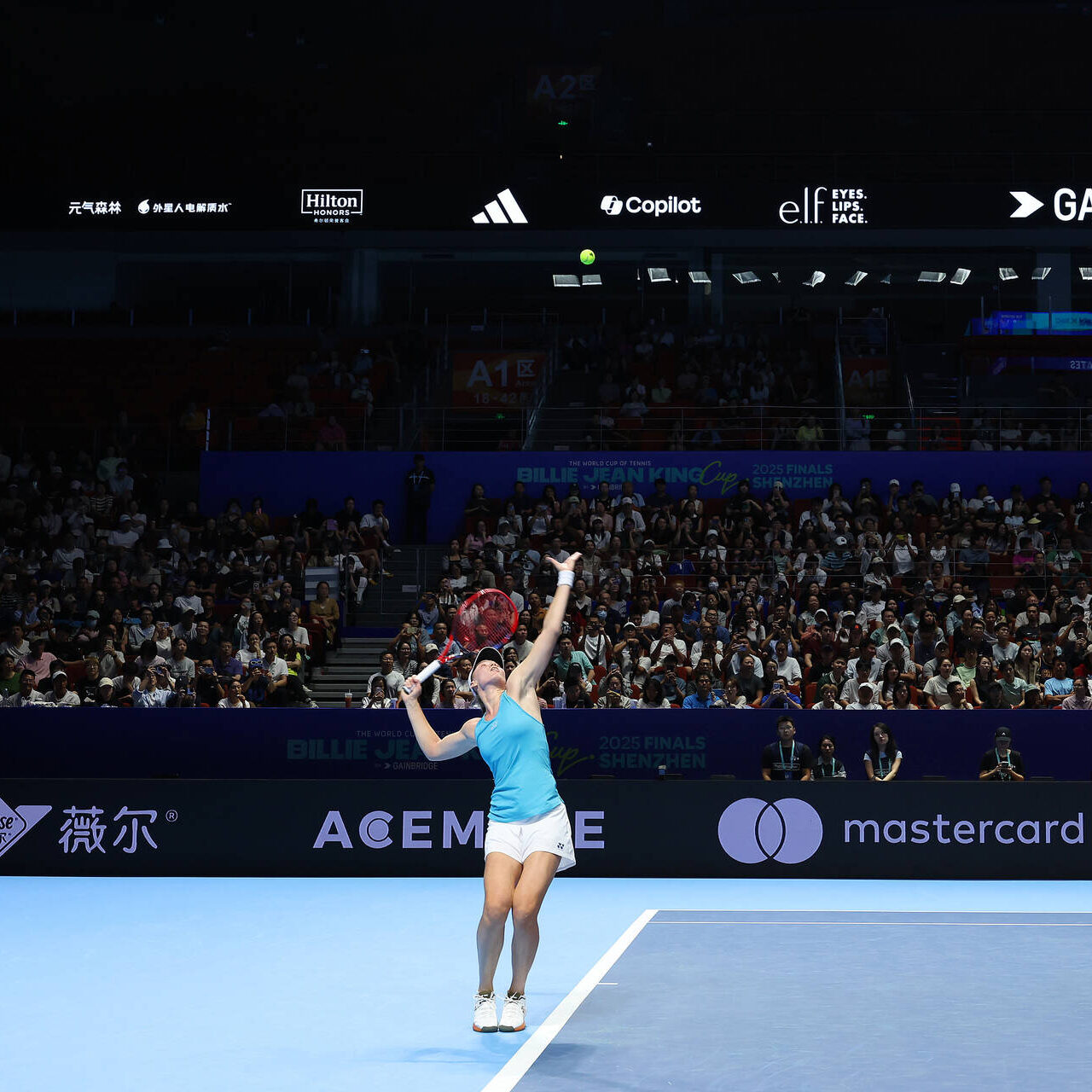 A tennis player in a blue top and white shorts reaches up to serve, racket raised, during the Billie Jean King Cup Finals 2025 in Shenzhen, China, with a packed