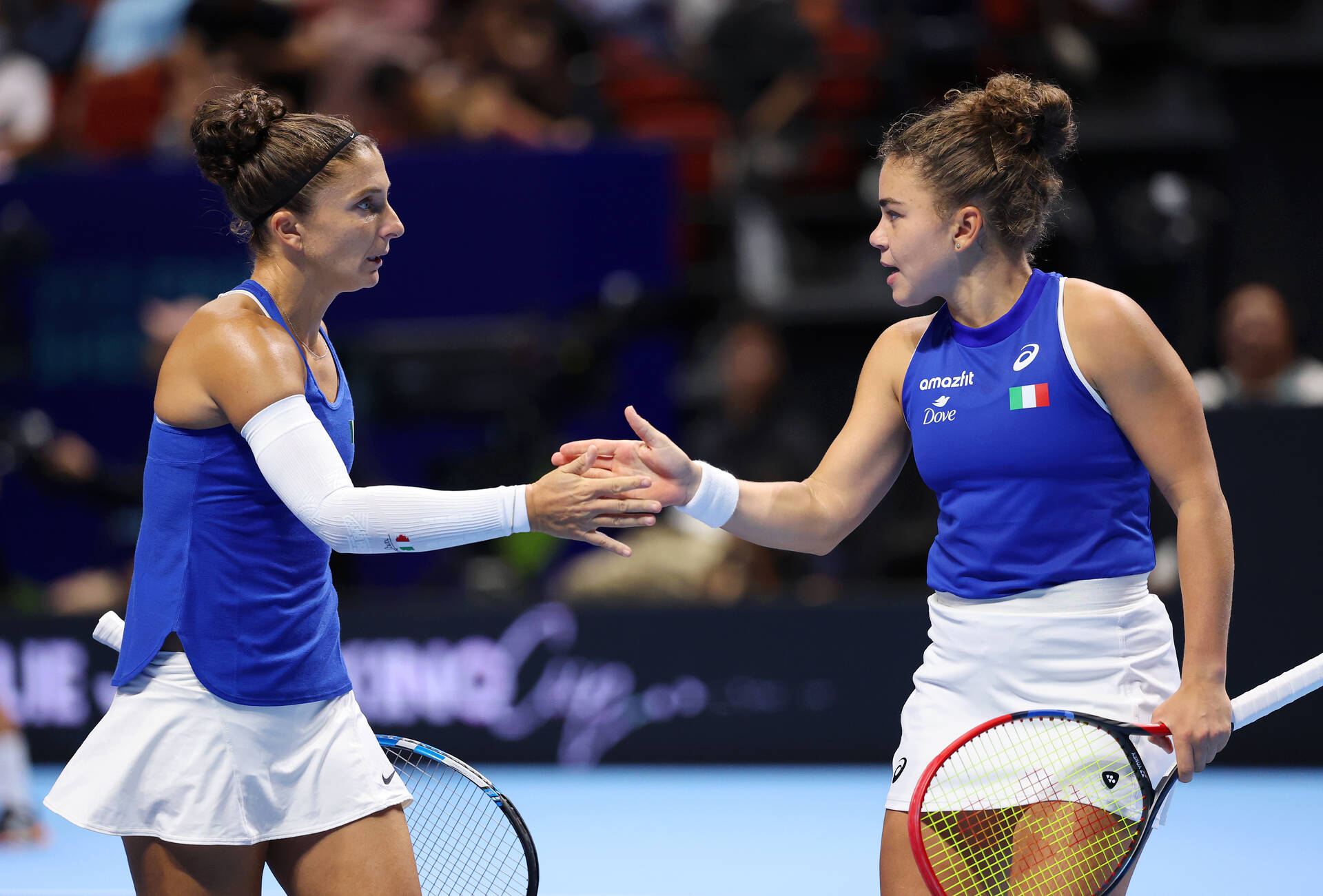 Two Italian tennis players in blue and white ASICS uniforms exchange a hand touch during a doubles match at the 2025 Billie Jean King Cup Finals, with Italian flag