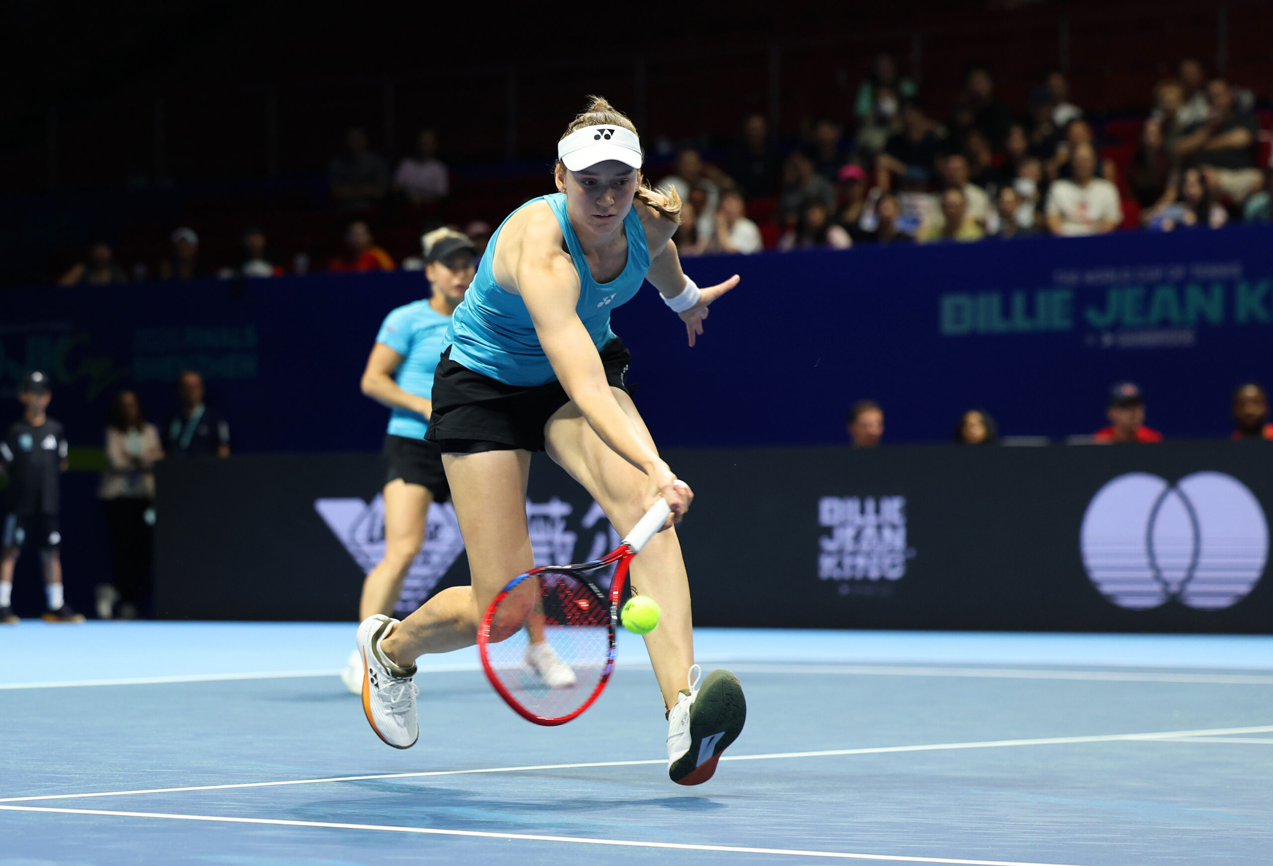 Female tennis player wearing a teal tank top, black shorts, and a white Yonex visor lunging to hit a backhand shot during the Billie Jean King Cup Finals 2025, with
