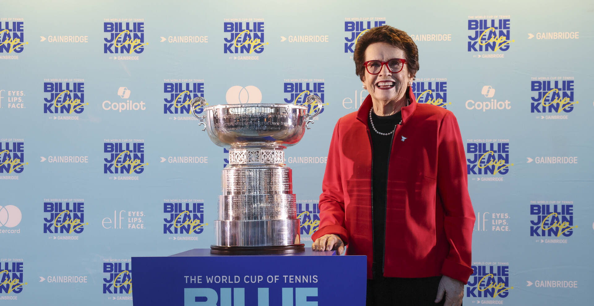 Smiling woman in a red jacket and red-framed glasses posing beside the Billie Jean King Cup trophy at the World Cup of Tennis Billie Jean King Cup by Gainbridge