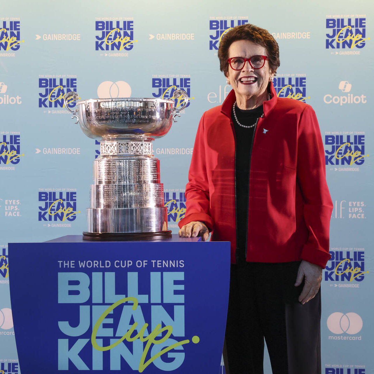 A smiling woman in a red jacket and red-framed glasses stands beside the Billie Jean King Cup trophy at a branded media backdrop for the Billie Jean King Cup by