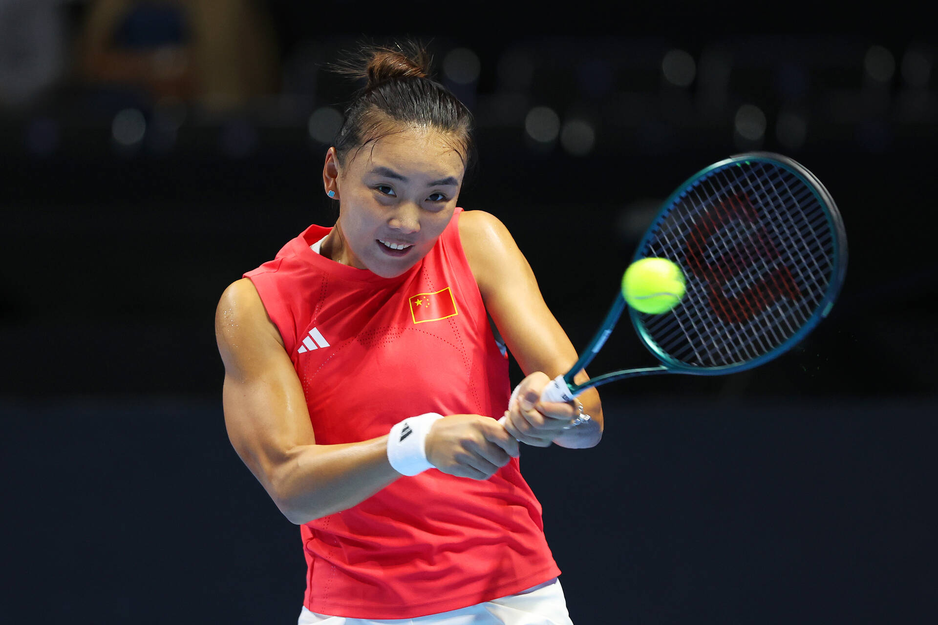 Chinese tennis player hitting a two-handed backhand during the Billie Jean King Cup Finals 2025 against Italy, wearing a red Adidas sleeveless top with the Chinese
