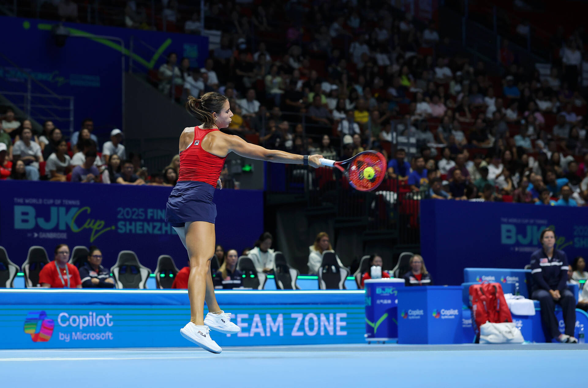 A female tennis player wearing a red Fila top and navy skirt leaps off the court to hit a forehand shot during the Billie Jean King Cup Finals 2025 in Shenzhen