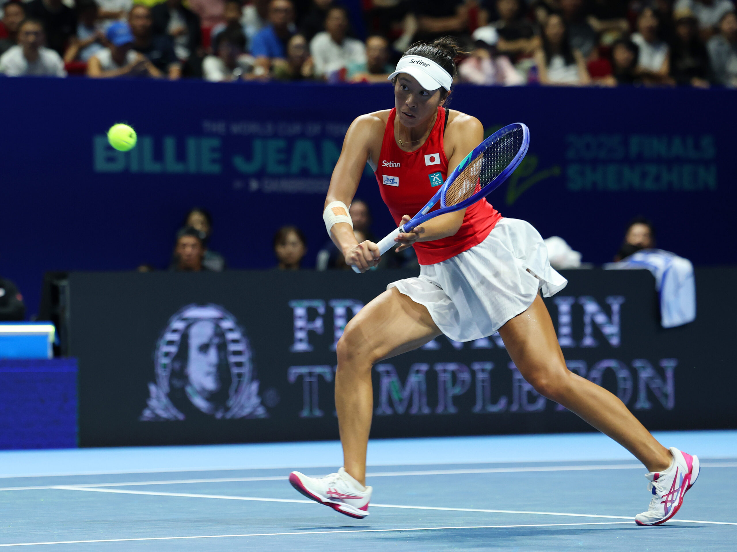 Japanese tennis player wearing a red kit with the Japanese flag patch, lunging to hit a backhand shot during the Billie Jean King Cup Finals 2025 in Shenzhen, with