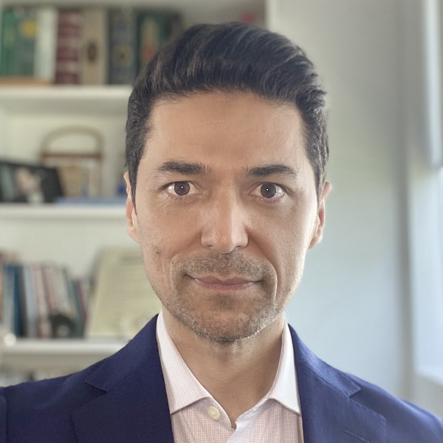 Professional headshot of a man with dark hair, wearing a navy blazer and light pink dress shirt, with a bookshelf visible in the background
